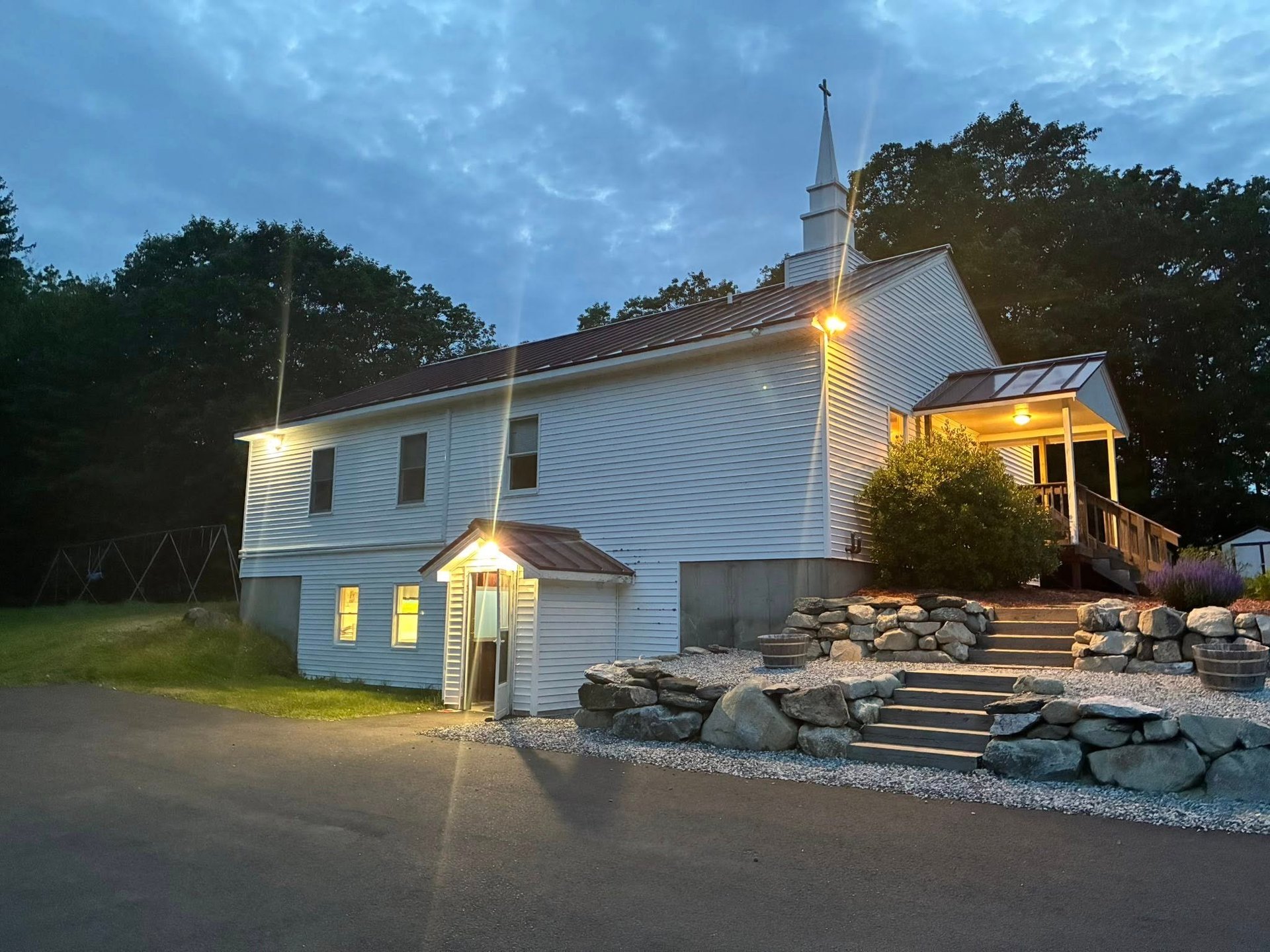 White country church illuminated at dusk with steeple, surrounded by landscaped rocks and trees